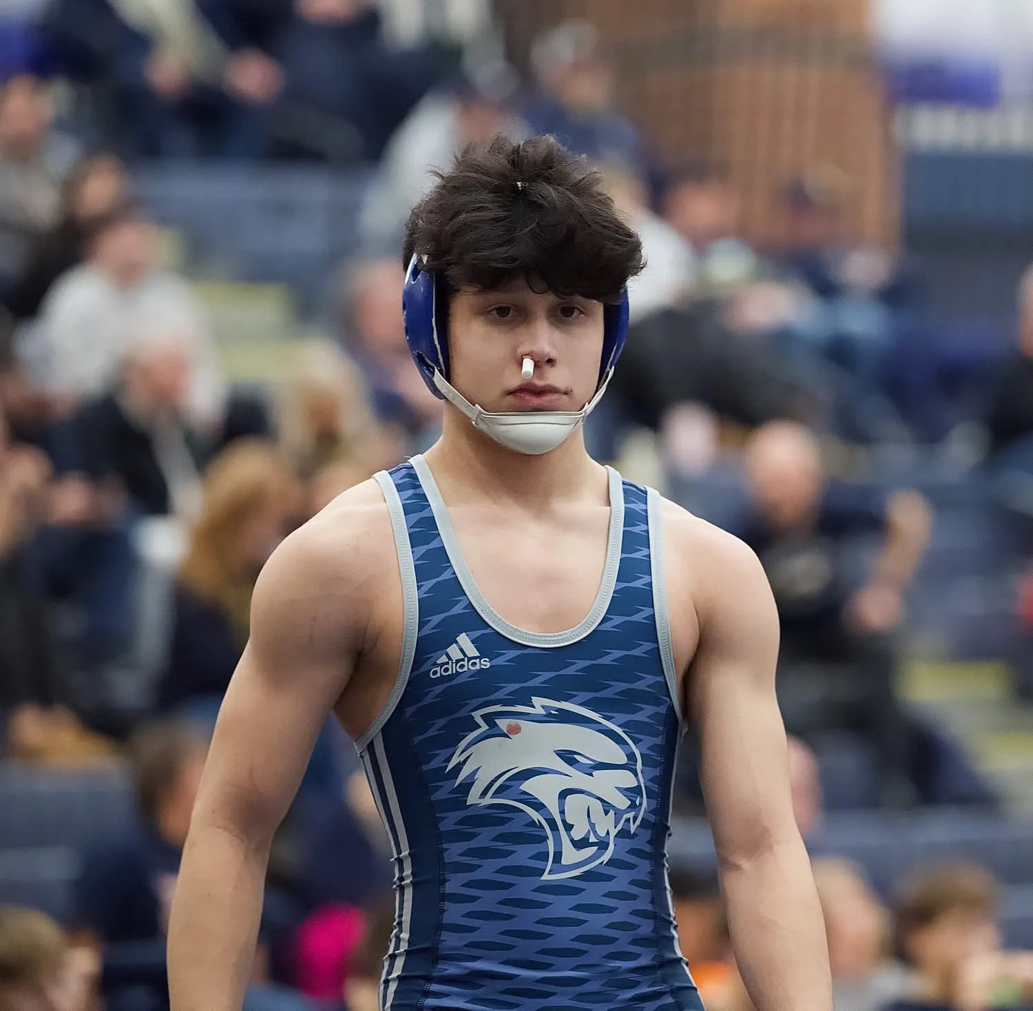 A wrestler in headgear waiting at the edge of the mat
